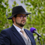 Mężczyzna w kapeluszu i okularach stoi przy mikrofonie z fioletową osłoną. Ma na sobie granatowy garnitur i beżowy krawat. W tle zielone liście.