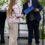 Dziewczynka w kwiecistej bluzie otrzymuje nagrodę od mężczyzny w kapeluszu. Tło to zielone krzewy i biały budynek. Scena na świeżym powietrzu.