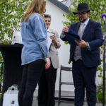 Na zdjęciu troje ludzi na scenie podczas ceremonii. Mężczyzna w garniturze i kapeluszu wręcza dokument kobiecie w dżinsowej kurtce. W tle zielone drzewa i biały budynek.