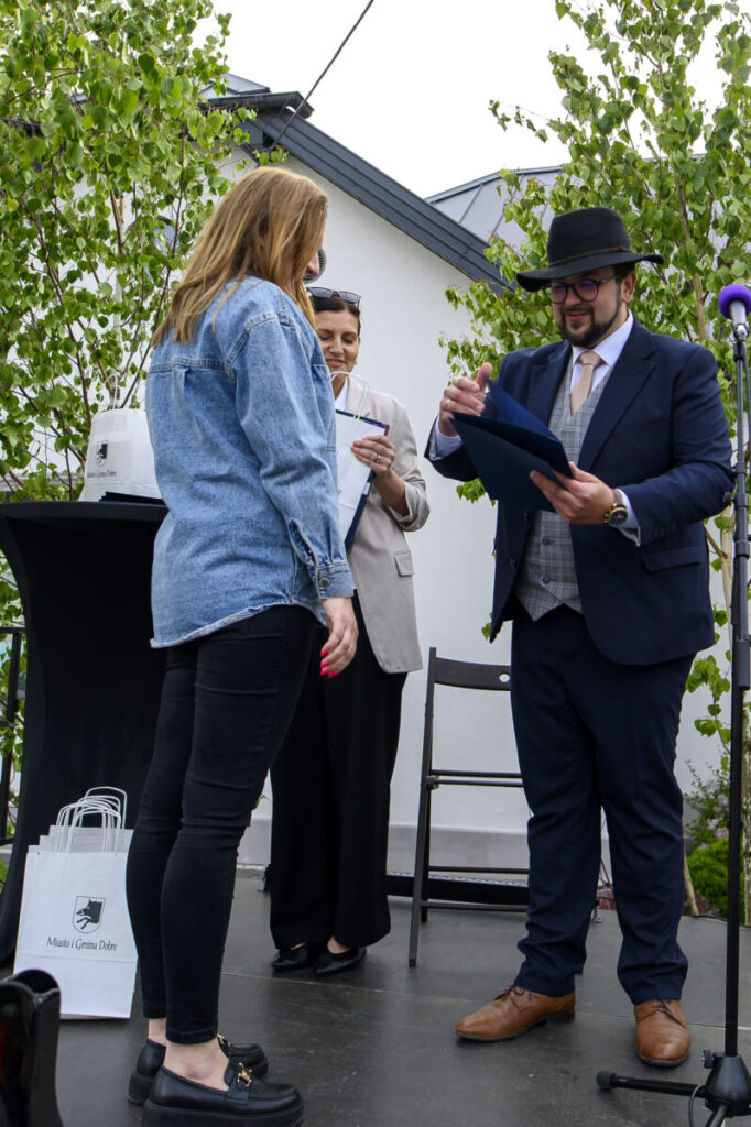Na zdjęciu troje ludzi na scenie podczas ceremonii. Mężczyzna w garniturze i kapeluszu wręcza dokument kobiecie w dżinsowej kurtce. W tle zielone drzewa i biały budynek.