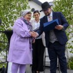 Trzy osoby na scenie, w tym kobieta w fioletowym płaszczu i mężczyzna w garniturze, trzymający certyfikat. W tle zielone drzewa. Mężczyzna w kapeluszu podaje dokument.