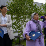 Trzy osoby stoją na zewnątrz, jedna mówi do mikrofonu. Noszą eleganckie ubrania w jasnych kolorach. W tle widać zielone krzewy.