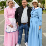 Trzy osoby na placu, dwie kobiety w kolorowych sukienkach retro z kapeluszami, mężczyzna w kurtce. Tło z drzewami.