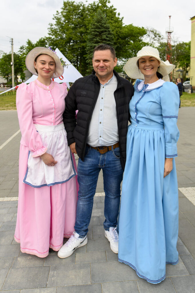 Trzy osoby na placu, dwie kobiety w kolorowych sukienkach retro z kapeluszami, mężczyzna w kurtce. Tło z drzewami.