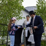 Dziewczynka otrzymuje książkę od elegancko ubranych dorosłych. W tle zielone drzewa, na pierwszym planie gitara.