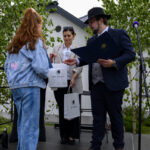 Na zdjęciu widać osobę wręczającą torby z napisem Miasto i Gmina Dobra. W tle biały budynek i zielone drzewa. Scena ma mikrofon po prawej stronie.