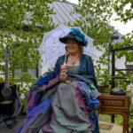 Kobieta w kolorowej sukni i kapeluszu z parasolką, siedzi na tarasie. Obok stoi gitara, w tle zielone drzewa i biały dom.