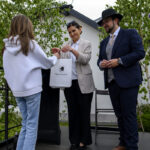 Trzy osoby stoją na scenie. Kobieta w jasnej marynarce wręcza torbę z logo Miasto i Gmina Dobre. Tło stanowi zielone drzewo.