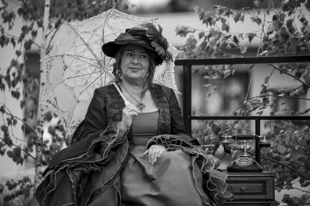 Kobieta w eleganckiej sukni z parasolką siedzi na zewnątrz obok retro telefonu. Tło to drzewa i ogrodzenie. Zdjęcie jest czarno-białe.