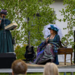 Dwie kobiety w strojach historycznych na scenie. Jedna trzyma książkę, druga siedzi z parasolką. Tło to zielone drzewa.