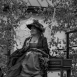 Kobieta w stroju retro z koronkowym parasolem siedzi przy stoliku z telefonem. W tle drzewa i budynek. Ujęcie czarno-białe.