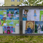 Tablica korkowa ze szkolnymi rysunkami o tematyce baśniowej, w jasnych i żywych kolorach. Tło zielone z żółtym budynkiem. Rysunki przedstawiają zamek, księżniczki i krajobrazy.