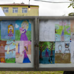 Tablica ogłoszeniowa z dziecięcymi rysunkami w jasnych kolorach. Obrazy przedstawiają postaci i krajobrazy, w tle żółty budynek. Obok rosną zielone gałęzie drzew.