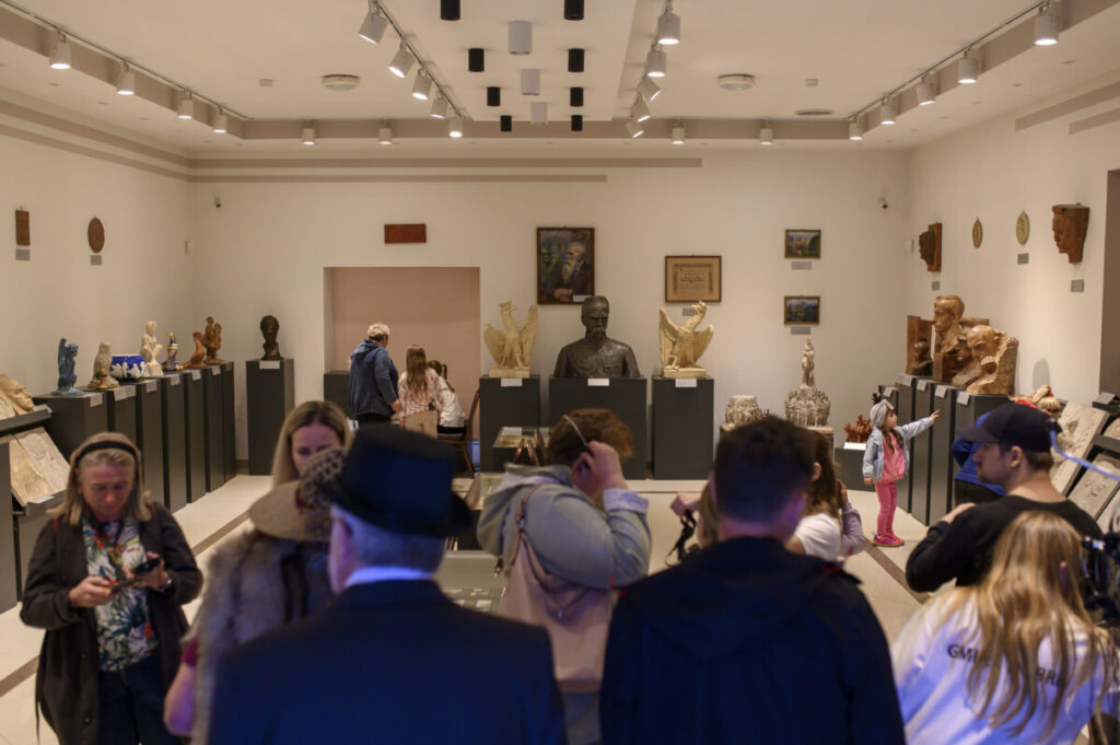 Na obrazie widoczna jest sala muzealna z rzeźbami i popiersiami. Ściany są jasne, a wystawa ma eleganckie oświetlenie. Kilka osób ogląda eksponaty w tle.