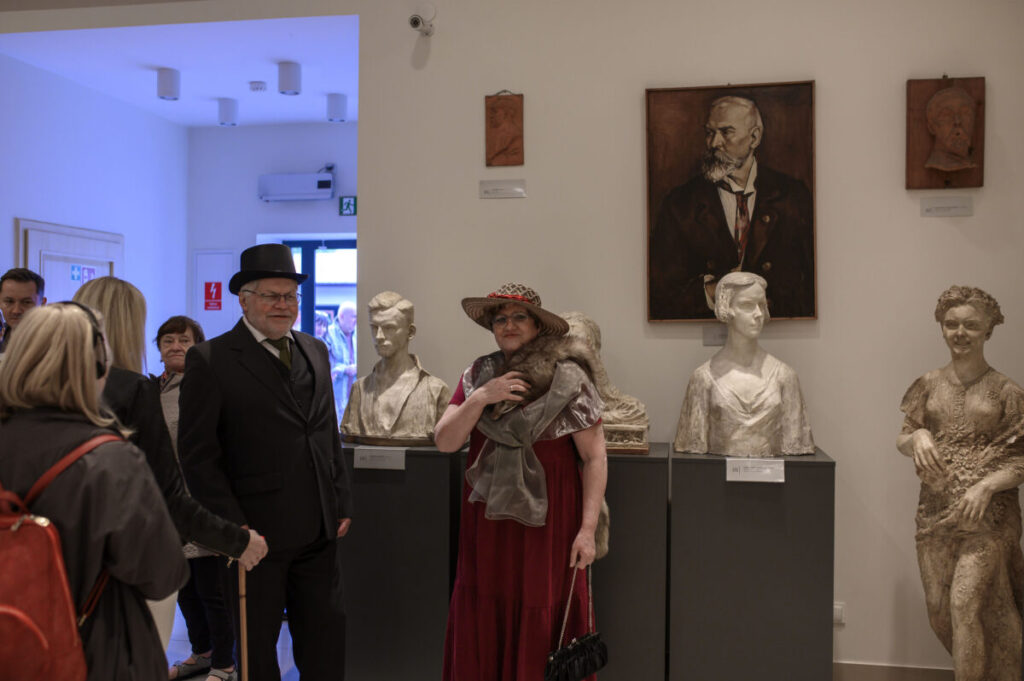 W muzeum, ludzie w strojach z epoki i rzeźby na postumentach. Dominuje biel ścian i kilka dzieł sztuki na tle. Atmosfera retro w eleganckim otoczeniu.