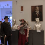 W muzeum, ludzie w strojach z epoki i rzeźby na postumentach. Dominuje biel ścian i kilka dzieł sztuki na tle. Atmosfera retro w eleganckim otoczeniu.