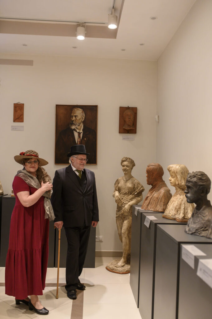 Dwie osoby w strojach z epoki stoją w muzeum. W tle popiersia i portret na jasnych ścianach. Oświetlenie punktowe.