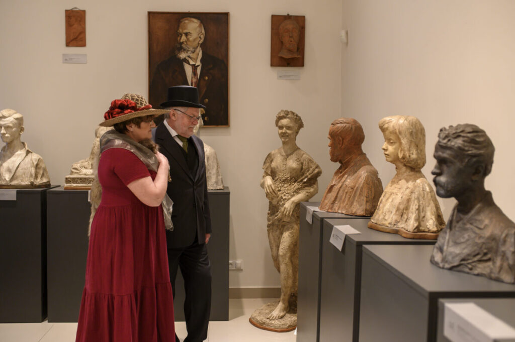 Muzeum z rzeźbami i popiersiami w różnych kolorach. Dwoje ludzi ubranych w stylu retro ogląda ekspozycję. Jasne ściany z portretami w tle.