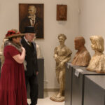 Muzeum z rzeźbami i popiersiami w różnych kolorach. Dwoje ludzi ubranych w stylu retro ogląda ekspozycję. Jasne ściany z portretami w tle.