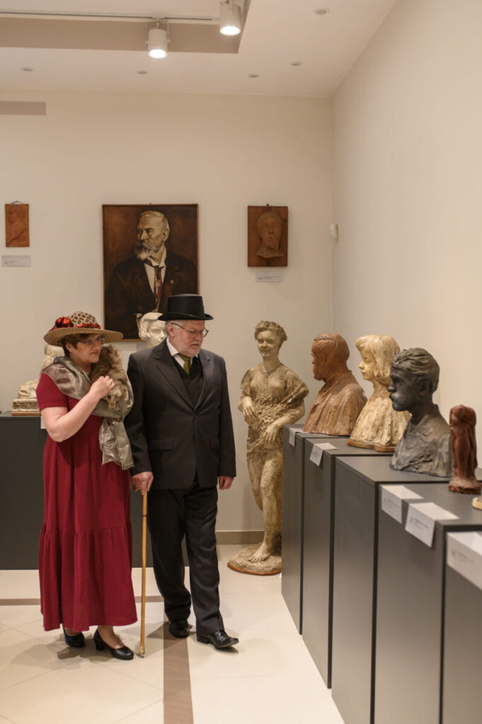 Dwójka ludzi w eleganckich strojach ogląda rzeźby w muzeum. Ściany są jasne z wiszącymi portretami. Tło jest minimalistyczne.