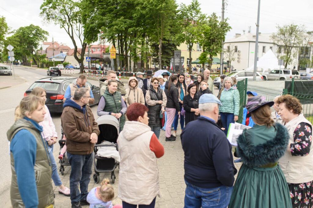Grupa ludzi stoi na chodniku w mieście; ubrani w różne kurtki, w tle drzewa i budynki. Kolorowe ubrania i zielone tło.