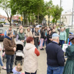 Grupa ludzi stoi na chodniku w mieście; ubrani w różne kurtki, w tle drzewa i budynki. Kolorowe ubrania i zielone tło.