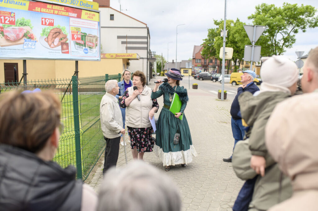 Grupa ludzi na chodniku, w tle billboard reklamowy. Kobieta w kostiumie z epoki trzyma mikrofon. Obok zielony płot i drzewa.
