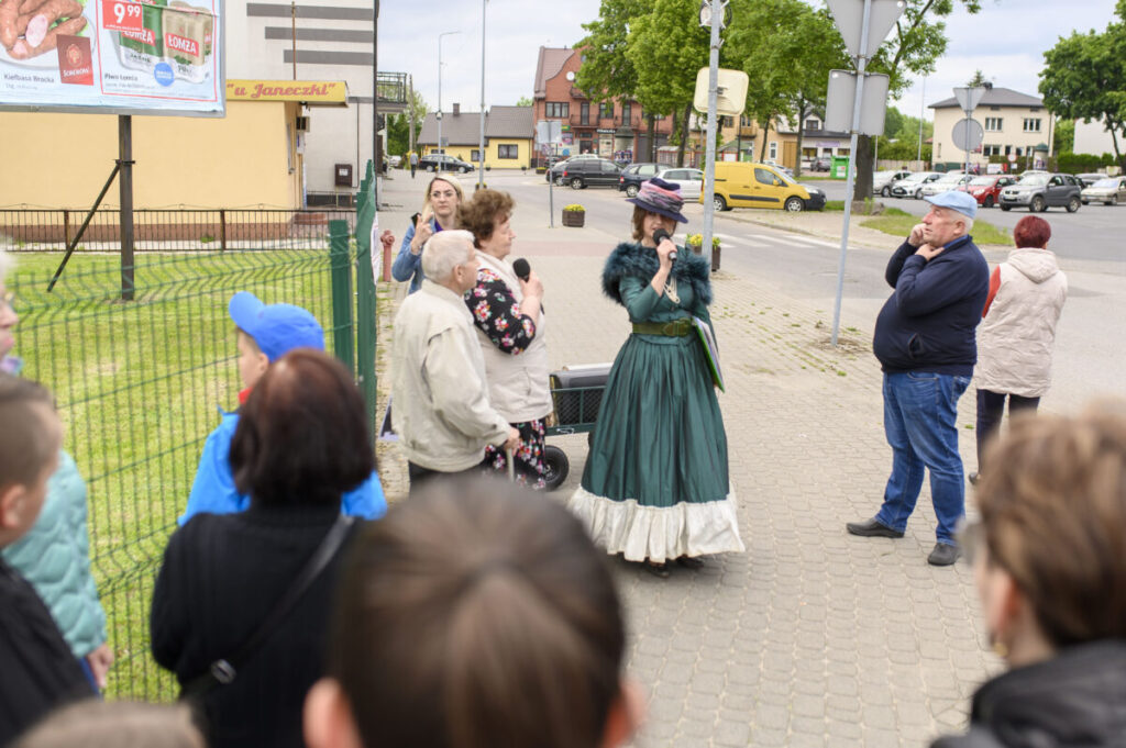 Grupa ludzi stoi na chodniku w mieście, jedna osoba w zielonej sukni mówi do mikrofonu. W tle budynki, samochody i zielone drzewa. Po lewej stronie widoczny jest billboard z reklamą.