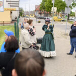 Grupa ludzi stoi na chodniku w mieście, jedna osoba w zielonej sukni mówi do mikrofonu. W tle budynki, samochody i zielone drzewa. Po lewej stronie widoczny jest billboard z reklamą.