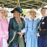 Cztery kobiety w strojach historycznych stoją pod zielonym namiotem z lampkami. Jedna ma różową suknię, inne w zieleni, błękicie i granacie. W tle stół z białymi dekoracjami.