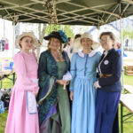 Cztery kobiety w kolorowych, historycznych strojach stoją pod zielonym namiotem ze światełkami. Każda ma na sobie kapelusz. Tło to zieleń trawy i inne osoby.