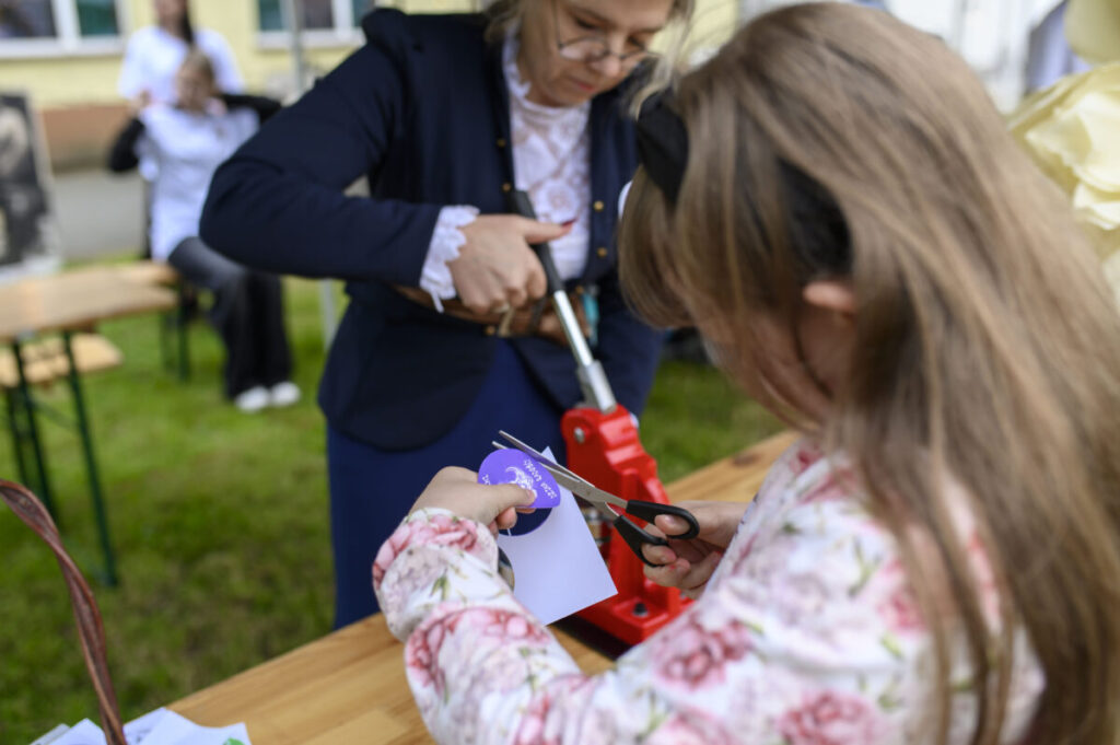 Dziewczynka w kwiecistej bluzce wycina fioletowy kształt. Obok stoi kobieta w ciemnej marynarce, trzymająca czerwone urządzenie. Tło jest rozmyte, na zewnątrz.