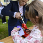 Dziewczynka w kwiecistej bluzce wycina fioletowy kształt. Obok stoi kobieta w ciemnej marynarce, trzymająca czerwone urządzenie. Tło jest rozmyte, na zewnątrz.