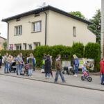 Na zdjęciu grupa osób stoi na chodniku przed kremowym budynkiem. W tle zielone krzewy. Dzieci są na rowerkach.