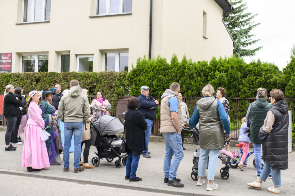 Grupa ludzi stoi przed beżowym budynkiem. Niektóre osoby są w kolorowych strojach, inne z wózkami. W tle zielone krzewy.