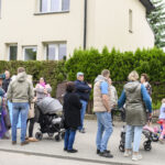 Grupa ludzi stoi przed beżowym budynkiem. Niektóre osoby są w kolorowych strojach, inne z wózkami. W tle zielone krzewy.