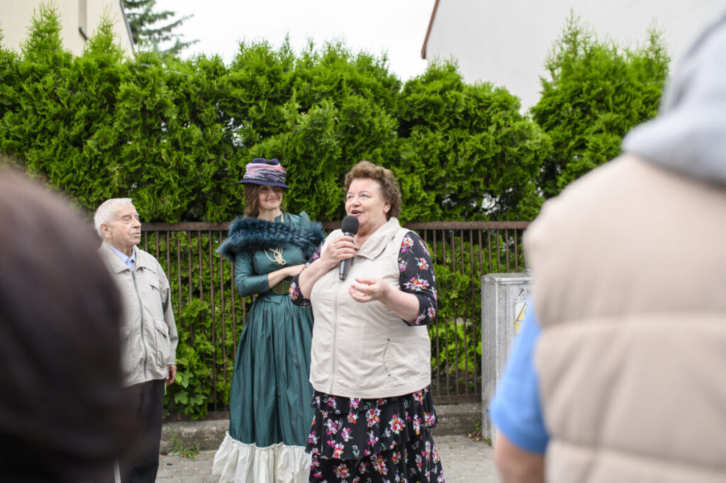 Trzy osoby w rozmowie na tle zielonych krzewów. Kobieta trzyma mikrofon, ubrana w kwiecistą sukienkę. Jedna z osób ma strój w stylu retro.