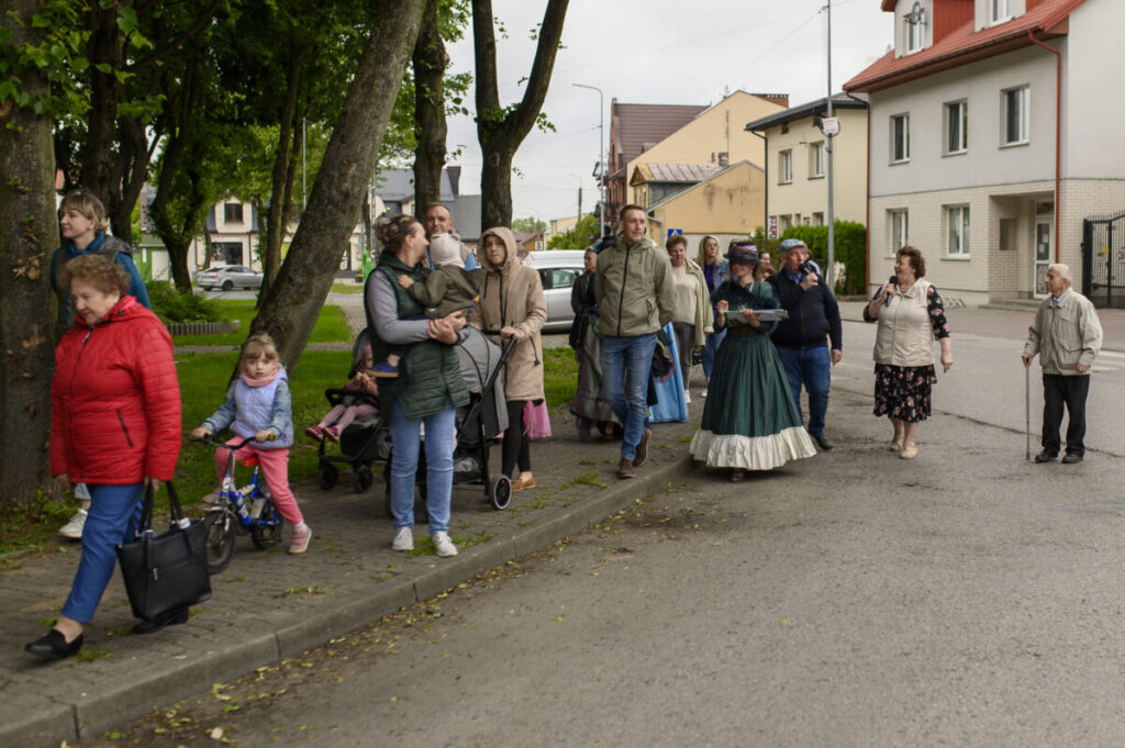 Grupa ludzi spaceruje po chodniku; tło z drzewami i domami. Osoby w różnym wieku, jedna osoba w historycznym stroju. Tło zielone i szare.