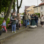 Grupa ludzi spaceruje po chodniku; tło z drzewami i domami. Osoby w różnym wieku, jedna osoba w historycznym stroju. Tło zielone i szare.