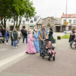 Grupa osób spaceruje po ulicy obok kamienic, część ubrana w historyczne stroje. Przewaga zieleni i pastelowych barw. Na trotuarze wózek z dzieckiem.