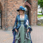 Osoba w zielonej sukni i kapeluszu z niebieskimi piórami stoi przed ceglanym murem. Tło z zielonymi drzewami. Uśmiechnięta postać trzyma chustkę.