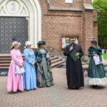 Grupa ludzi w historycznych strojach stoi przed ceglaną budowlą z dużymi, ozdobnymi drzwiami. Kolorowe suknie dominują pośród zieleni drzew. Tło stanowi czerwono-ceglana ściana i zielony trawnik.