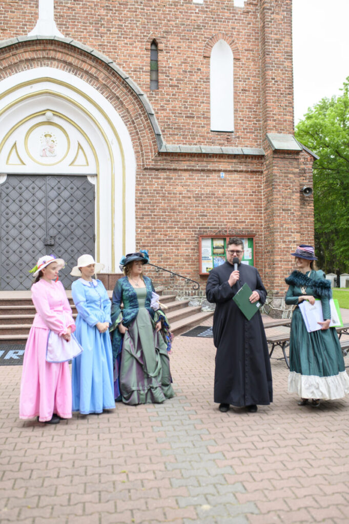 Grupa osób w kolorowych strojach historycznych stoi przed ceglaną fasadą kościoła. Na środku mężczyzna w czarnym stroju, trzyma mikrofon. Tło stanowi drzwi kościoła i zieleń po prawej stronie.