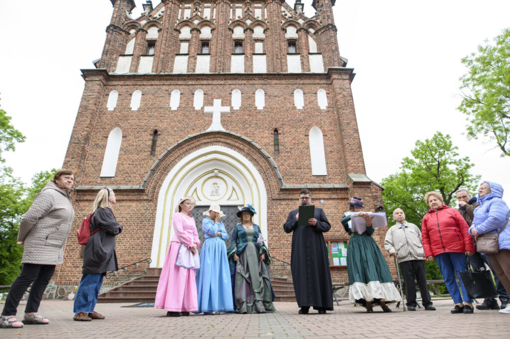 Grupa ludzi przed ceglaną katedrą. W centrum osoby w historycznych strojach. Tło zielone drzewa i biało-czerwony budynek.
