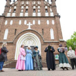 Grupa ludzi przed ceglaną katedrą. W centrum osoby w historycznych strojach. Tło zielone drzewa i biało-czerwony budynek.