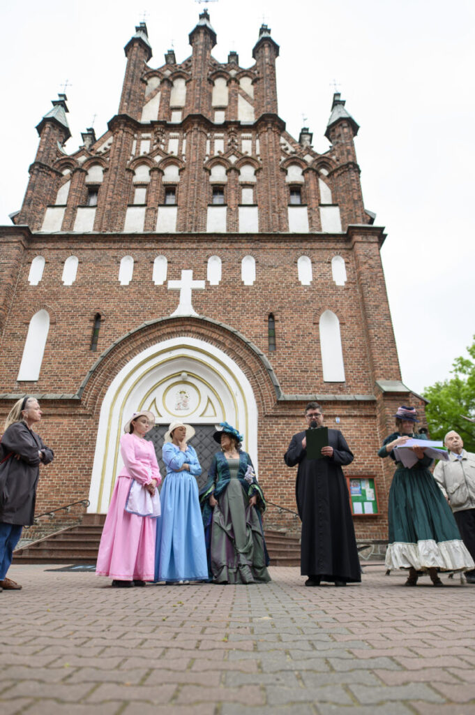 Kościół z cegły, ludzie w strojach historycznych przed wejściem. Jasne kolory sukien. Tło zielone drzewa.