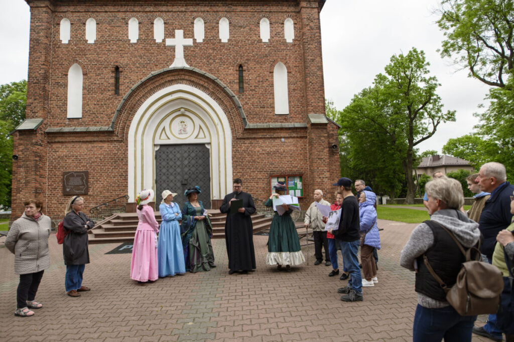 Grupa ludzi w strojach historycznych stoi przed ceglanym kościołem z białymi detalami. Przewodnik w czarnej szacie przemawia. W tle zielone drzewa.