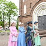 Cztery kobiety w kolorowych, historycznych strojach stoją przed ceglaną fasadą kościoła. Kolory sukni to różowy, niebieski, zielony. Tło to drzewa i brukowana nawierzchnia.