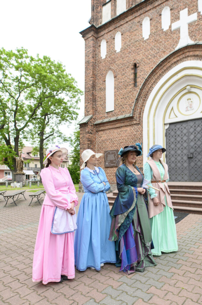 Cztery kobiety w kolorowych, historycznych strojach stoją przed ceglaną fasadą kościoła. Kolory sukni to różowy, niebieski, zielony. Tło to drzewa i brukowana nawierzchnia.