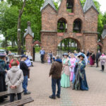 Grupa ludzi w kolorowych strojach historycznych stoi przed ceglanym budynkiem z dzwonami. Drzewa zielenią się w tle. Na pierwszym planie tętni życie społeczne.
